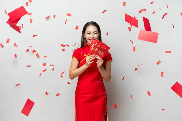 Smiling happy Asian woman in traditional oriental costume holding red envelopes Ang Pow in gray background, Chinese text means great luck great profit and and everything goes well