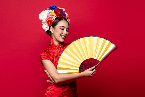 Smiling Asian woman wearing traditional dress with flower chaplet holding golden fan in isolated studio red background for Chinese new year concept