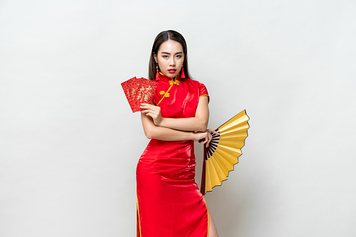 Asian woman in oriental cheongsam costume holding red envelopes Ang Pow and fan posing on light gray studio background for Chinese new year concepts, foreign texts mean great luck great profit