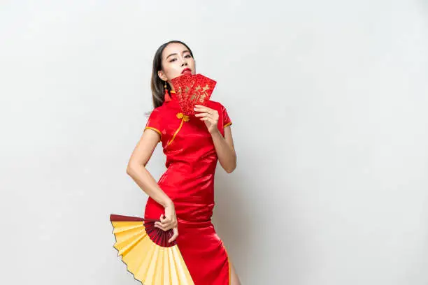 Asian woman in oriental cheongsam costume holding red envelopes Ang Pow and fan posing on light gray studio background for Chinese new year concepts, foreign texts mean great luck great profit