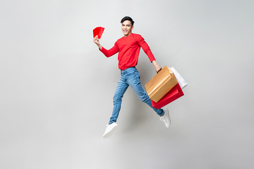 Surprised Asian man holding bags and red envelope Ang Pow   jumping in isolated light gray studio background for Chinese new year shopping concept