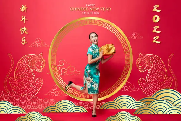 Happy Asian woman in traditional cheongsam dress holding gold package on red oriental background for 2022 year of the tiger, foreign text translates as Chinese new year
