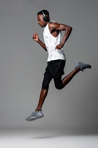 Full length muscular African sportsman in sportswear and headphones listening to music and running during workout against gray background