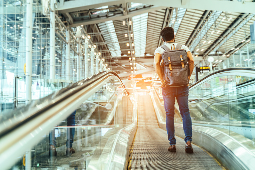 New normal and Travel insurance concept.Young caucasian man wearing face mask and carrying backpack on escalator or travelator walkway in airport terminal,protection during coronavirus pandemic.