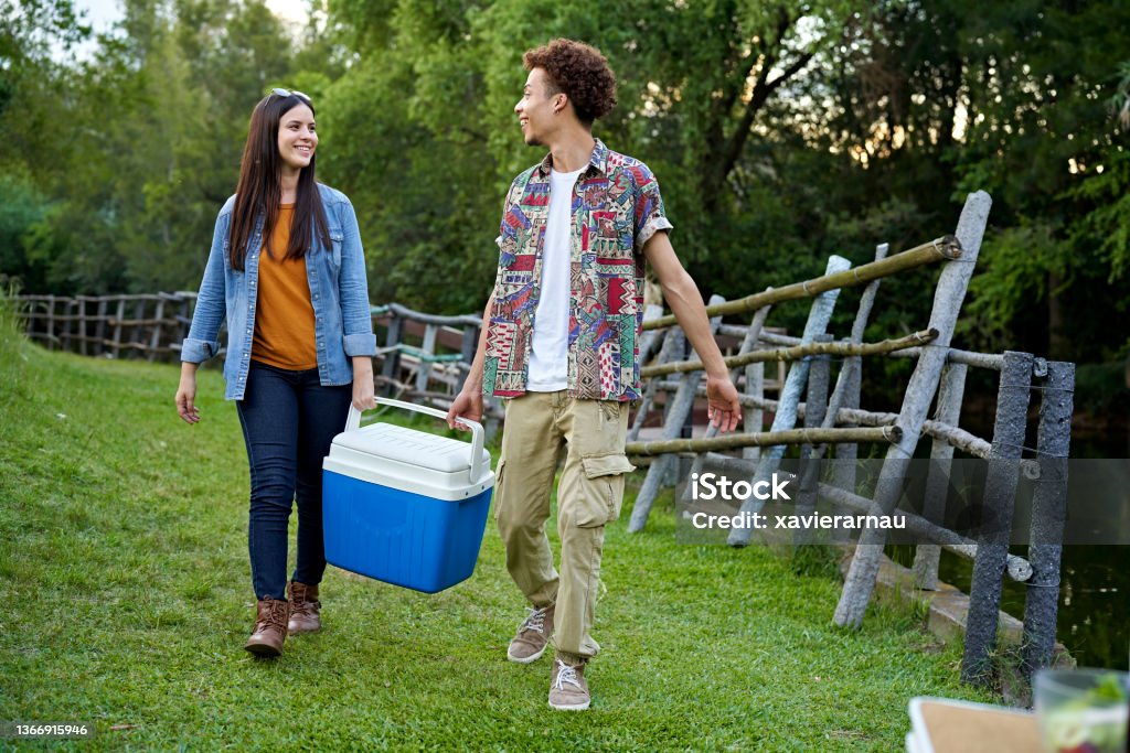 Young friends carrying cooler while camping in woodland area Full length front view of Hispanic woman and African-American man in casual clothing smiling and talking as they approach picnic table. Cooler - Container Stock Photo