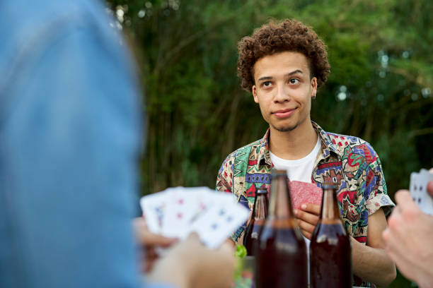 Young man enjoying beer and cards outdoors with friends Waist-up view with focus on African-American man in casual clothing holding playing cards and looking across table at opponent with uncertain expression. smirking stock pictures, royalty-free photos & images