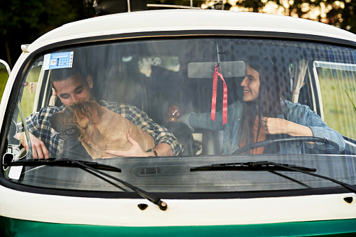 Front view of man and woman in casual clothing photographed through van windshield as they sit in front seat with Shar-Pei while traveling.