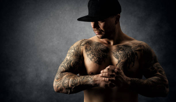 Bodybuilder with Tattoo. Strong Muscular Athletic Body Man. Power Athlete in Hat over Dark Grunge Artistic Studio Background. Mysterious Handsome Tattooed Men Portrait Bodybuilder with Tattoo. Strong Muscular Athletic Body Man. Power Athlete in Hat over Dark Gray Grunge Artistic Studio Background. Mysterious Handsome Tattooed Men Portrait Hiding Face man muscular chest stock pictures, royalty-free photos & images