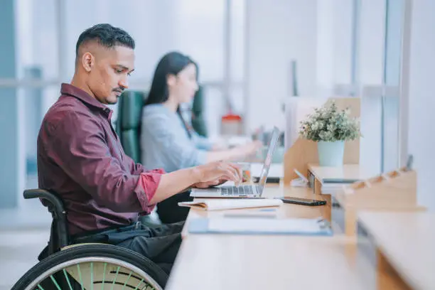 Photo of Asian Indian white collar male worker in wheelchair concentrating working in office beside his colleague