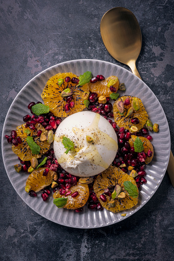 Recipe of; mandarin oranges, burrata, pistachios and pomegranate. Burrata cheese is recognisable  by its loose texture: small, soft cheese curds and cream packed inside an outer shell like mozzarella. Colour, vertical format with some copy space.