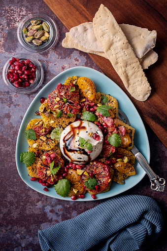 Recipe of; mandarin and blood oranges, burrata, pistachios and pomegranate seeds. served with crispy flatbreads.\nBurrata cheese is recognisable  by its loose texture: small, soft cheese curds and cream packed inside an outer shell like mozzarella. Colour, vertical format with some copy space.