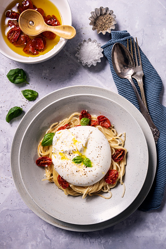Close-up view of a recipe of freshly cooked spaghetti with a pesto and olive oil with some oven dried tomatoes. Burrata cheese is recognisable  by its loose texture: small, soft cheese curds and cream packed inside an outer shell like mozzarella. Colour, vertical format with some copy space.