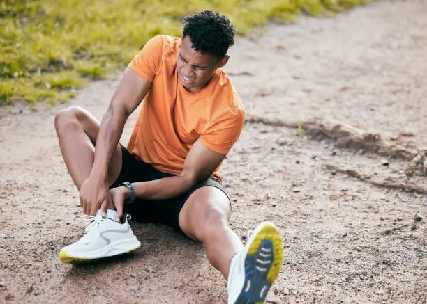 Photo of Full length shot of a young man sitting alone outside and suffering from an ankle injury during his workout