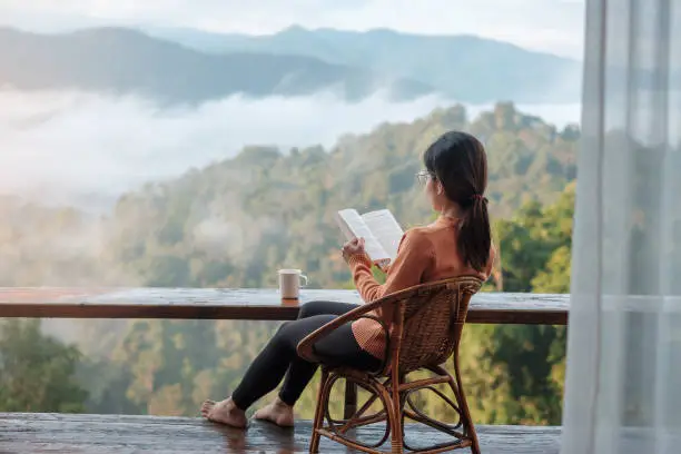 Photo of young woman reading book near window and looking mountain view at countryside homestay in the morning sunrise. SoloTravel, journey, trip and relaxing concept