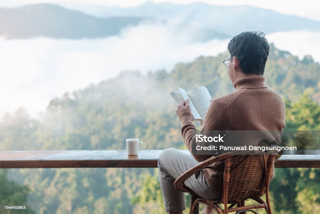 young man reading book near window and looking mountain view at countryside homestay in the morning sunrise. SoloTravel, journey, trip and relaxing concept Window Stock Photo