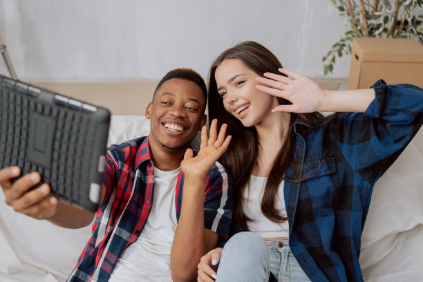istock Young smiling couple sits on couch secured with foil dark skinned handsome man holds tablet in hands, they are having video conversation wave to their parents from their new renovated apartment 1366938231