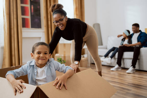 mom playing with baby, pushing a cardboard box across the living room floor where her sweet little son sits, joy at moving into a new apartment, smiling boy - family large american culture fun imagens e fotografias de stock