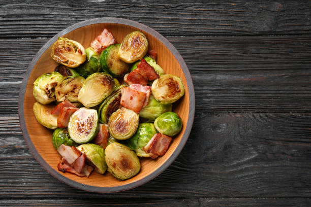 istock Delicious fried Brussels sprouts with bacon in bowl on wooden table, top view. Space for text 1366940723