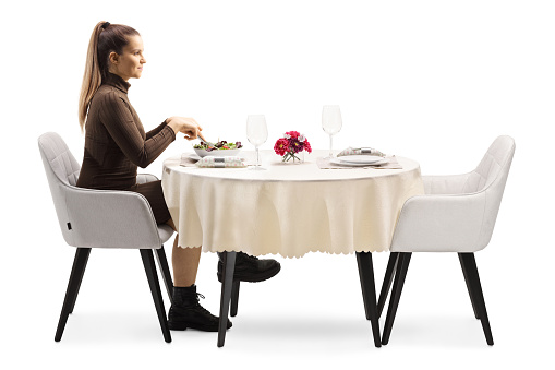 Profile shot of a young woman sitting at a restaurant table and eating a salad isolated on white background