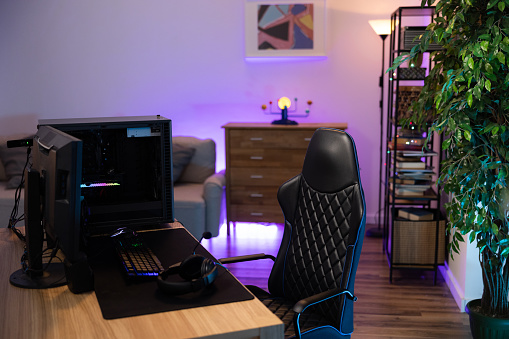 A backlit professional computer for a gamer. The monitor stands on a desk. The cozy room with modern design is lit with purple blue neon led lights.