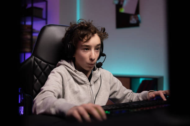 istock Focused young boy with curly hair spends time at computer playing multiplayer video games with team, talks to people through headset, dark room night lit by neon led lights 1366941657