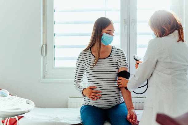 Doctor with a pregnant woman wearing medical masks during an examinations Pregnant woman and female doctor with face masks during examination in hospital. Doctor is measuring pressure to her patient women health clinic for pregnant women stock pictures, royalty-free photos & images