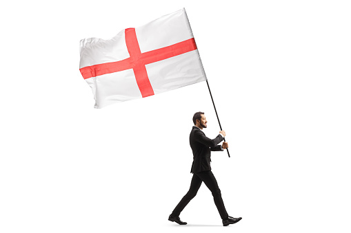Full length profile shot of an elegant man in a suit walking and carrying the flag of England isolated on white background