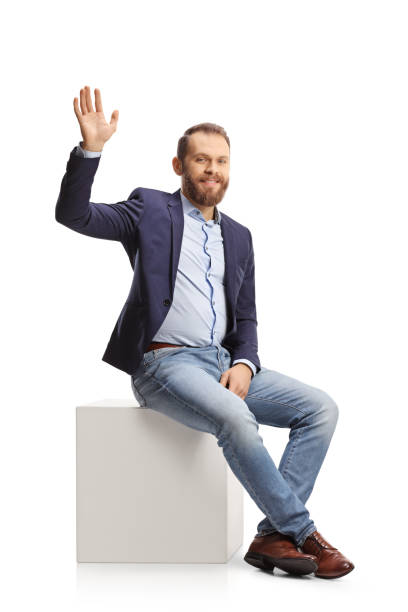 istock Young man in suit and jeans sitting on a white cube and waving 1366943546