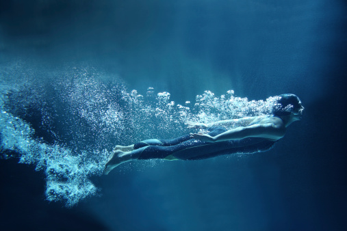 Athlete is dressed in a professional black swimwear. She is swimming horizontally like she was flying. She has two hands together along the body. She is looking ahead.  Behind her body you can see a lot of air bubbles. The background is dark blue- like open water.