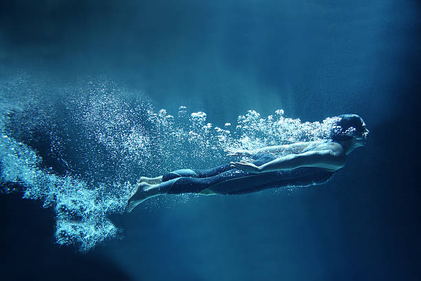 Female swimmer underwater flowing on blue background Athlete is dressed in a professional black swimwear. She is swimming horizontally like she was flying. She has two hands together along the body. She is looking ahead.  Behind her body you can see a lot of air bubbles. The background is dark blue- like open water. underwater diving stock pictures, royalty-free photos & images