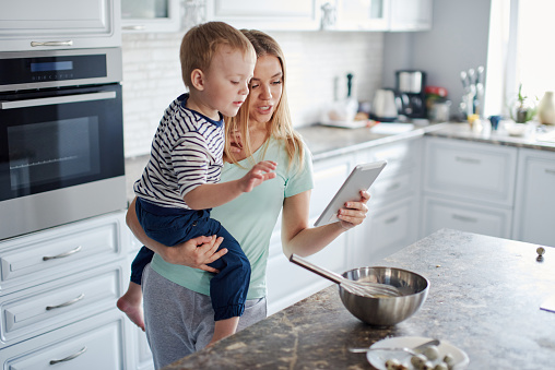 Portrait of young caucasian blond woman with her son using tablet at the kitchen together. Busy mom is holding son and tablet. Stay connected, social network, stay home concept