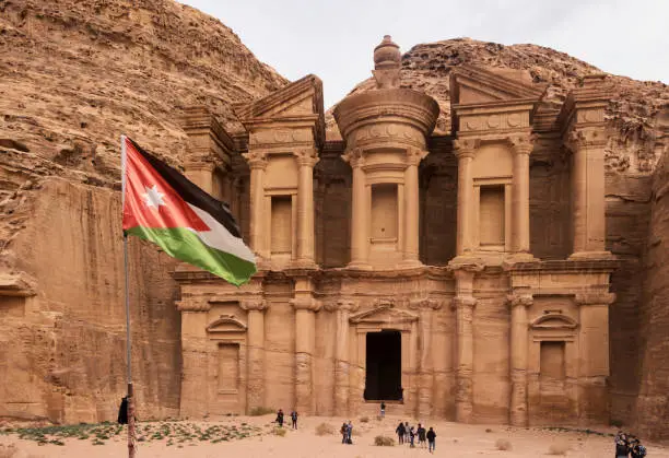 Jordan - Petra - The tourists visiting Ad Deir (the monastery), built in 3 BCE as nabataean tomb, now is one of most famous monuments in archeological park with jordanian national flag on a foreground