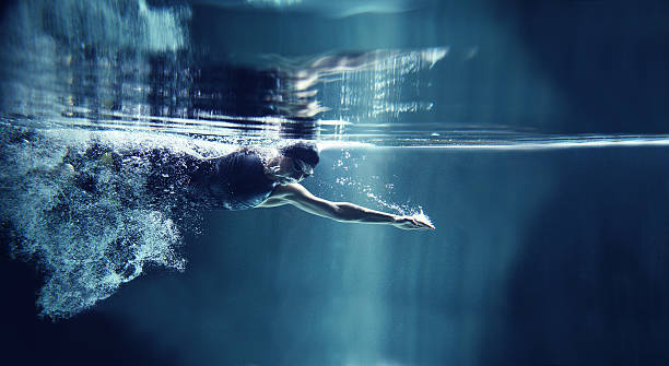 Athlete swimming freestyle on blue background, underwater view A female athlete is swimming crawl. She is wearing professional, black swim wear, swimming glasses and cap. You can see her torso, head and one hand. She is emerging from air bubbles. She is exhaling air to water and has one arm streatched in front. She is looking down. In the top of image you can see the surface of water and reflection of the scene. The background is dark blue. There are no swimming pool elements. This is a horizontal image.  muscle photos stock pictures, royalty-free photos & images