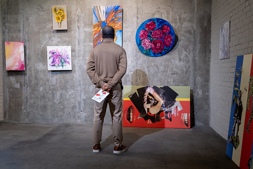 Rear view of African man standing in front of the wall with paintings and looking at them in art gallery