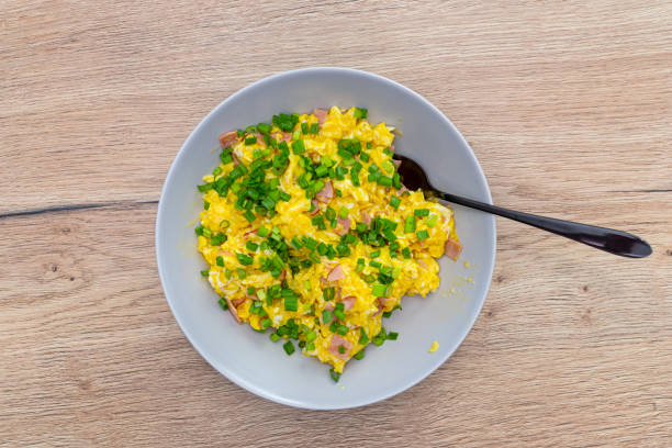 Scrambled eggs with chives and pieces of ham, lying on a plate with a fork on a wooden table, top view.