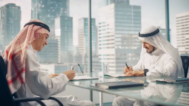 Photo of Two Happy Emirati Businessmen in White Traditional Kandura Sitting in Office and Signing Contract. Business Partners Sign Lucrative Investment. Saudi, Emirati, Arab Businessman Concept.