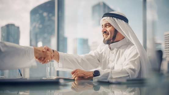 Smiling Emirati Businessman in White Traditional Kandura Sitting in Office with Business Partners. Shake Hands on Successful Investment. Saudi, Emirati, Arab Businessman Concept.