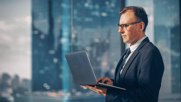 istock Confident Businessman in a Suit Standing in Modern Office, Using Laptop Computer, Looking out of the Window on Big City with Skyscrapers. Successful Finance Manager Planning Work Projects. 1366951653