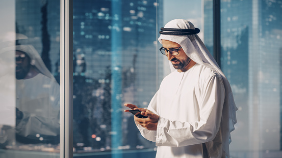 Exitoso hombre de negocios musulmán con atuendo blanco tradicional de pie en su oficina moderna, usando un teléfono inteligente junto a la ventana con rascacielos. Exitoso concepto de empresario saudí, emiratí y árabe. photo