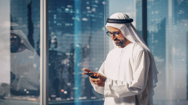 uomo d'affari musulmano di successo in abito bianco tradizionale in piedi nel suo ufficio moderno, usando lo smartphone accanto alla finestra con i grattacieli. concetto di successo saudita, emiratino, uomo d'affari arabo. - agal foto e immagini stock
