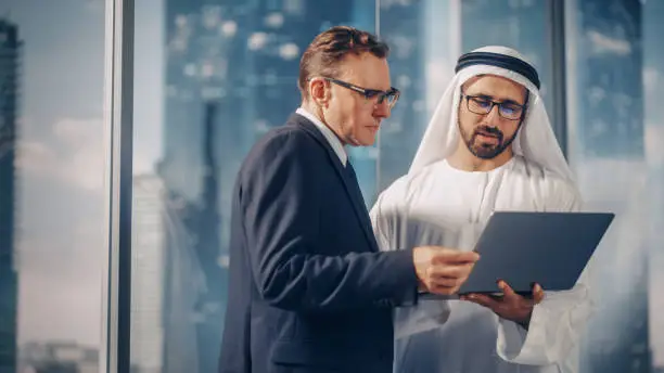 International Operations Manager Meeting Saudi Business Partner in Traditional Kandura. They're Standing in Modern Office, Using Laptop Computer. Successful Saudi, Emirati, Arab Businessman Concept.