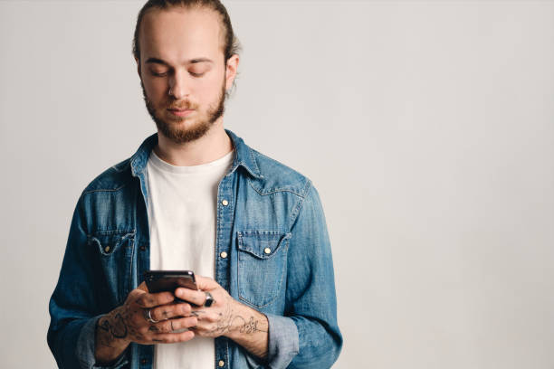 istock Attractive tattooed guy dressed in denim shirt texting with friends using smartphone isolated on white background. Modern technology 1366954766