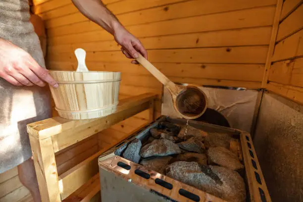 Close-up shot of unrecognizable man pouring water from wooden barrel on hot rocks in Finnish sauna.