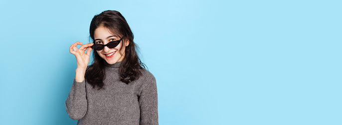 Style, flirt. Excited young beautiful girl wearing gray pullover in sunglasses isolated on blue background. Concept of emotions, facial expression, youth, inspiration, sales. Copy space for ad. Flyer