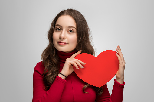 Beautiful girl in a red sweater is holding red cardboard heart. She is looking at the camera, smiling.