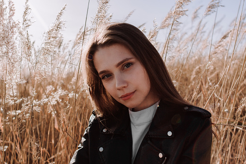 Smiling young woman in black leather jacket sitting in field of pampas grass in front of sky and sun. Urban style and street fashion. Girl in casual outfit looking at camera. Golden hour