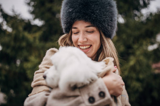 Beautiful woman hugging her Maltese dog in front of pine tree on a winter day outdoors.