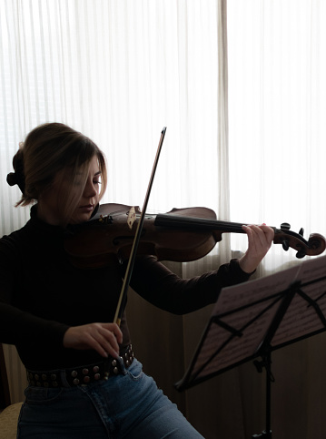Beautiful and young girl enjoying at her home playing her viola, practicing and enjoing her free time, playing near the window on the sunny day.
