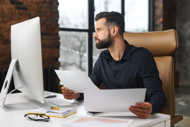 istock Clever and concentrated caucasian guy is doing paperwork. Man looking through paper sitting at the desk with a computer in office. Man checking report 1366966161