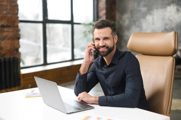 istock Smiling caucasian guy sitting at the office desk, using trendy laptop for a work. Young man is talking on smartphone and smiling to the camera, while having conversation with clients or colleagues 1366966627